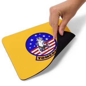 May include: A yellow mouse mat with a blue and red circle featuring a cartoon cat wearing a belt and holding a gun. The circle has white stars and the text "TOMCAT" in black.