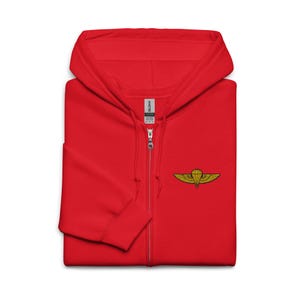 May include: A bright red zip-up hoodie with a hood and a small embroidered emblem on the chest. The emblem features a parachute design with gold-colored wings. The hoodie has a full-length zipper and a soft, comfortable appearance.