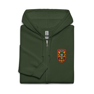 May include: A dark green zip-up hooded sweatshirt with a patch on the chest. The patch features a skull, wings, and the text "MAC SOG" in yellow and red. The sweatshirt has a full zipper and a hood.