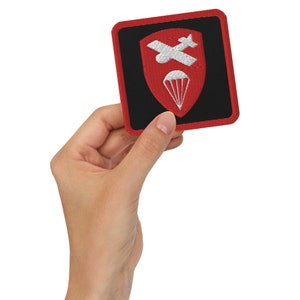 Può includere: Una toppa ricamata rossa e nera con un design di paracadute e aereo bianco. La toppa è circondata da un bordo rosso.
