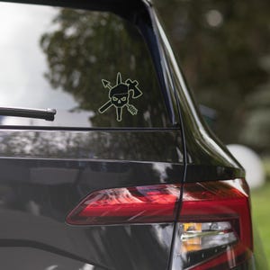 May include: A black and white sticker of a skull wearing a beret with a crossed axe and arrow behind it. The sticker is on the back window of a black car.