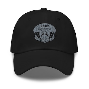 Puede incluir: Gorra de béisbol negra con el emblema de USAF Pararescue bordado. El emblema incluye un paracaídas, un águila y el texto "USAF Pararescue" y "That Others May Live". La gorra tiene una visera curva y una correa ajustable.