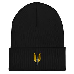 May include: Black beanie hat with a gold and silver embroidered emblem. The emblem is a winged design with a sword. The hat is made of a soft, knitted material.