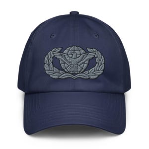 May include: A navy blue baseball cap featuring a centered, embroidered grey emblem of an eagle with wings spread wide, positioned in front of a globe and surrounded by a laurel wreath. The cap has a structured crown and a curved brim.