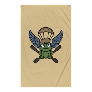 May include: A tan rectangular wall flag featuring a central graphic of a green frog wearing a tan beret. The frog is positioned beneath a parachute and between two blue wings, with two crossed dark brown oars displayed behind the frog's body.