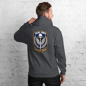 Può includere: Felpa con cappuccio grigio scuro con un emblema "Task Force Phantom Talon" sul retro. L'emblema presenta un teschio, una spada e ali in un design blu e oro. La felpa ha un cappuccio e una vestibilità rilassata.
