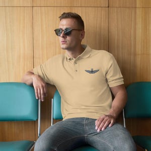 May include: A light tan polo shirt with a small, dark emblem on the chest. The shirt has short sleeves and a button-down collar. The person is wearing sunglasses and jeans.