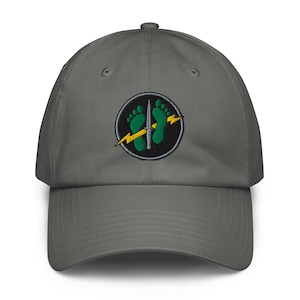 May include: A charcoal gray baseball cap featuring an embroidered circular patch on the front. The patch displays two green footprints, a silver vertical dagger, and a yellow lightning bolt set against a black background with a gray border.