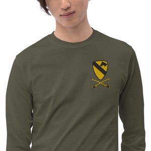 May include: Olive green long-sleeve shirt featuring an embroidered emblem of a yellow shield with a black diagonal stripe, a black axe, and crossed sabers. The shirt is made of a soft, comfortable material.