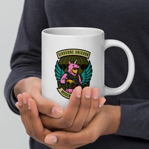 以下が含まれることがあります： 白いセラミックマグカップ。ピンクのコスチュームを着てライフルを持っているユニコーンのカラフルなグラフィックが特徴です。「AIRBORNE UNICORN」と「MODER」の文字が含まれています。マグカップには持ちやすい大きなハンドルが付いています。