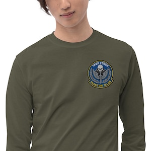 Könnte beinhalten: Olivgrünes Langarmshirt mit einem kreisförmigen Aufnäher auf der linken Brust. Der Aufnäher zeigt einen Totenkopf, Flügel und den Text "TASK FORCE PHANTOM TALON" in Blau und Gold.