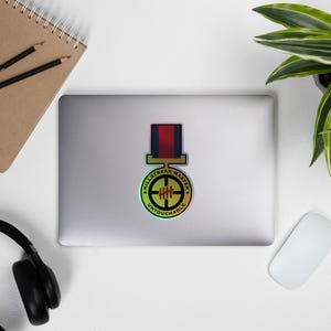 Puede incluir: Una pegatina holográfica con un diseño circular. El texto dice "KILLSTREAK MASTER UNTOUCHABLE". El diseño central es una mira con cuatro líneas horizontales. La pegatina tiene un esquema de color rojo, azul y dorado.