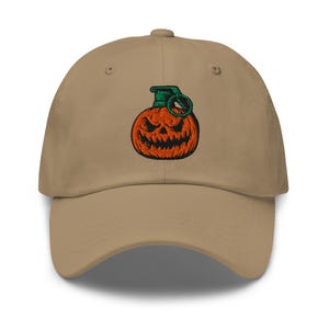 May include: A beige baseball cap featuring an embroidered design of a jack-o'-lantern grenade. The pumpkin is orange with a scary face, and the grenade pin is green. The cap has a curved peak and a button on top.
