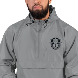 Könnte beinhalten: Graue Anorakjacke mit Kapuze und halbem Reißverschluss. Die Jacke hat ein schwarzes Emblem auf der Brust mit dem Schriftzug "DE OPPRESSO LIBER" in einem kreisförmigen Design. Die Jacke ist aus leichtem Material.