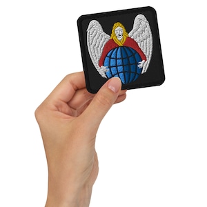 Puede incluir: Parche bordado que representa un ángel con alas blancas, una túnica roja y cabello rubio, sosteniendo un globo azul. El parche es cuadrado con un borde negro y está sostenido en una mano.