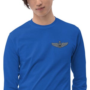 May include: A royal blue long-sleeve shirt featuring a silver-colored emblem on the upper left chest. The emblem depicts a star above a winged design. The shirt is made of a soft material.
