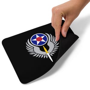 Könnte beinhalten: Schwarzes rechteckiges Mauspad mit einem kreisförmigen Emblem mit einem blauen und roten Stern, umgeben von weißen Flügeln und einem gelb-weißen Dolch. Das Mauspad wird von einer Hand hochgehalten.