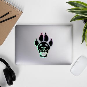 May include: A black and white sticker of a stylized paw print with a skull and teeth inside. The sticker is on a silver laptop.