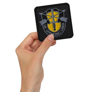 Puede incluir: Un parche bordado negro con un emblema amarillo y gris. El emblema presenta una espada, flechas y el texto "DE OPPRESSO LIBER".