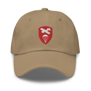 Può includere: Un cappello da baseball color beige con una toppa rossa ricamata che rappresenta un paracadute bianco e aerei incrociati. La toppa è un'insegna militare.