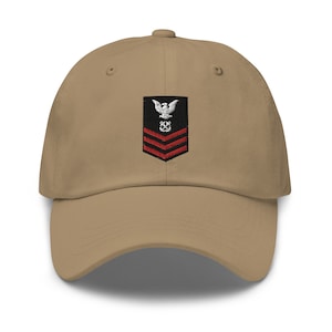 May include: A tan baseball cap with an embroidered patch featuring a white eagle, red stripes, and a black anchor. The patch is centered on the front of the cap.