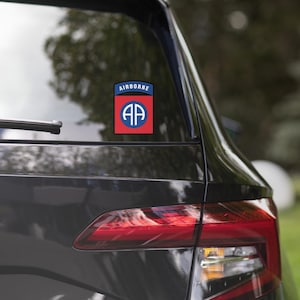 May include: A black car with a red, white, and blue "AIRBORNE" sticker on the back window. The sticker features a blue circle with the letters "AA" inside, surrounded by a red ring.