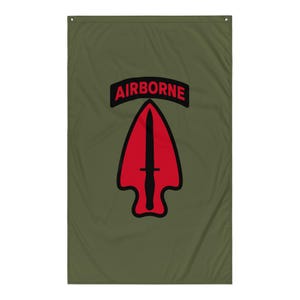 Könnte beinhalten: Olivgrüne Flagge mit rotem Pfeil-Design und schwarzem Dolch, mit dem Wort "AIRBORNE" in Rot. Die Flagge ist rechteckig und hat oben eine Öse zum Aufhängen. Das Design ist mittig auf der Flagge platziert.