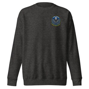 Könnte beinhalten: Dunkelgraues Sweatshirt mit Rundhalsausschnitt. Auf der linken Brust befindet sich ein kreisförmiger Aufnäher mit einem Totenkopf, Flügeln und den Worten "TASK FORCE" und "PHANTOM TALON" in blau-goldenem Design.