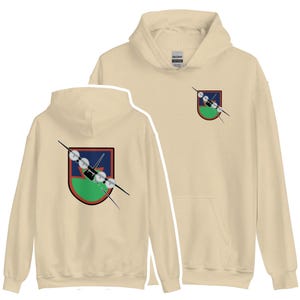 Puede incluir: Una sudadera con capucha beige con un parche verde, rojo y azul en la parte delantera y trasera. El parche presenta una silueta negra de un avión con una hélice.