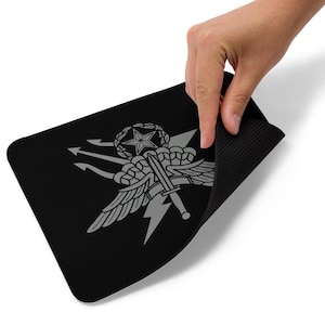 Könnte beinhalten: Schwarzes rechteckiges Mousepad mit einem grauen Emblem, das einen Stern, Flügel, einen Dreizack und ein Schwert zeigt. Das Mousepad wird angehoben, wodurch eine strukturierte schwarze Unterseite sichtbar wird. Das Emblem ist mittig platziert.