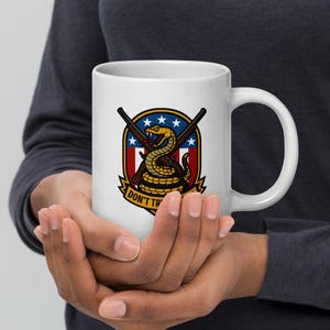 May include: White ceramic mug featuring a graphic of a coiled yellow snake with a red tongue, crossed rifles, and a patriotic shield design. The mug has the text "DON'T TREAD" in a banner. The mug is held by a person.