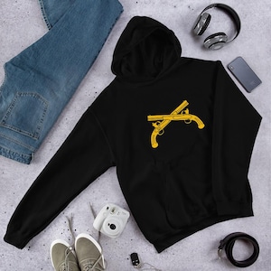 May include: Black hoodie with a gold crossed pistol graphic. The hoodie has a hood and a front pocket. The image also shows blue jeans, gray sneakers, a camera, headphones, a phone, and a black belt.