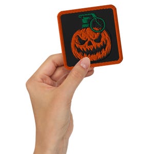 May include: Embroidered patch featuring a jack-o'-lantern grenade design. The patch has an orange pumpkin with a carved face, a green top, and a black background. The patch is square with an orange border.