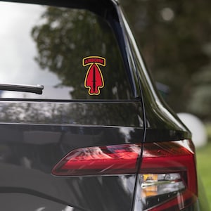 May include: A red and black Airborne sticker with a yellow border is attached to the rear window of a black car. The sticker shows a red arrowhead with a black sword and the word "AIRBORNE" in yellow.