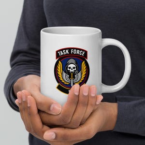 Op de afbeelding: Witte keramische mok met een Task Force-embleem. Het embleem toont een gehoodde schedel, een zwaard en vleugels, met de woorden "TASK FORCE" in een rode banner. De mok wordt vastgehouden.