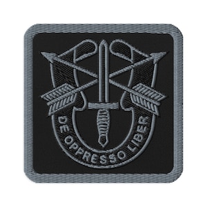 May include: Embroidered patch featuring a black background with a gray border. The design includes a central dagger, two crossed arrows, and the Latin phrase "DE OPPRESSO LIBER" in a curved banner. The patch has a square shape.