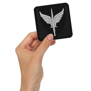 Pode incluir: Um remendo bordado preto com um design de espada alada branca. O remendo é quadrado e tem uma borda preta.