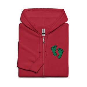 Könnte beinhalten: Ein roter Kapuzenpullover mit durchgehendem Reißverschluss. Der Hoodie hat zwei grüne Fußabdrücke auf der Vorderseite. Der Markenname "Gildan" ist auf einem Etikett in der Nähe des Reißverschlusses zu sehen.