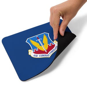 May include: A blue rectangular mousepad with the Air Combat Command emblem. The emblem features a sword, wings, and the text "AIR COMBAT COMMAND". The mousepad has a black, textured underside.