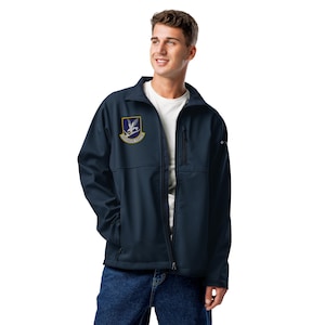 May include: A navy blue softshell jacket featuring a white t-shirt underneath, a vertical zippered chest pocket, and a patch on the right chest displaying an eagle emblem with the text Defensor Fortis. The jacket is worn over blue denim jeans.