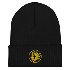 May include: A black cuffed beanie featuring an embroidered yellow circular patch on the front. The patch displays a stylized design of a vertical sword positioned next to a wing, all enclosed within a thin yellow circular border on a black background.
