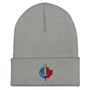 May include: A heather gray knit beanie featuring a cuffed brim. Centered on the cuff is an embroidered patch depicting a split circular design: the left side shows a blue and green globe, the right side a red maple leaf, bisected by a silver sword.