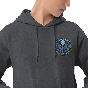 Könnte beinhalten: Dunkelgrauer Kapuzenpullover mit Kordelzug. Auf der Brust befindet sich ein kreisförmiger Aufnäher mit dem Text "TASK FORCE PHANTOM TALON" um einen Totenkopf, Flügel und ein Schwertdesign. Der Aufnäher ist blau, gelb und weiß.
