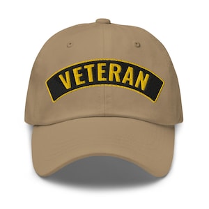 May include: A tan baseball cap with a black patch that says "Veteran" in yellow lettering.