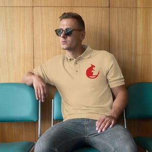 Może przedstawiać: Beżowa koszulka polo z czerwonym logo kangura na lewej piersi. Koszulka ma klasyczny kołnierzyk i zapięcie na guziki. Osoba nosi okulary przeciwsłoneczne i dżinsy.