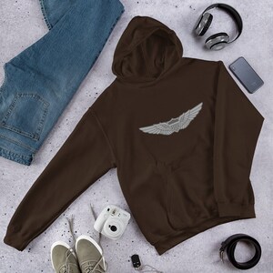 Pode incluir: Um moletom marrom com um emblema de asas branco no peito. A imagem inclui jeans azuis, tênis cinza, fones de ouvido, um smartphone, uma câmera e um cinto preto. O moletom tem capuz e um bolso frontal.