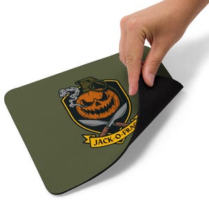 May include: An olive green rectangular mousepad with a Halloween design. The graphic features a jack-o'-lantern with a grenade top, crossed knives, and the text "JACK-O-FRAG" on a banner. The mousepad has a black, textured underside.