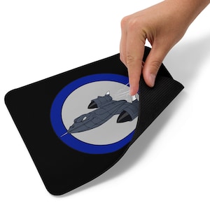 Może przedstawiać: Czarna podkładka pod mysz z niebieskim kółkiem i kreskówkowym obrazkiem czarnego samolotu SR-71 Blackbird w locie.