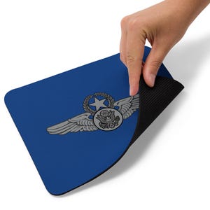 May include: Blue rectangular mousepad featuring a silver emblem with wings, a star, and a circular design. The emblem is centered on the mousepad. The mousepad has a black, textured underside. A hand is lifting the edge of the mousepad.