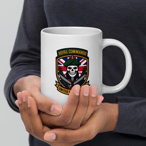 Op de afbeelding: Witte keramische mok met een Royal Commando-embleem. Het embleem toont een schedel met baret, gekruiste messen en een Britse vlag. De tekst "ROYAL COMMANDO" en "UNITED" zijn ook zichtbaar.